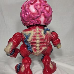 Puede incluir: Una figura anat&oacute;mica roja y blanca con una cabeza en forma de cerebro. La figura tiene detalles de m&uacute;sculos y huesos expuestos, con acentos azules. El juguete est&aacute; sobre una superficie blanca.