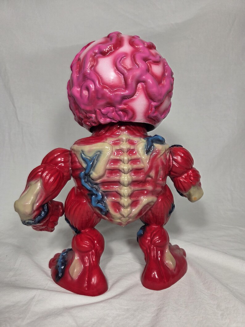 Puede incluir: Una figura anat&oacute;mica roja y blanca con una cabeza en forma de cerebro. La figura tiene detalles de m&uacute;sculos y huesos expuestos, con acentos azules. El juguete est&aacute; sobre una superficie blanca.