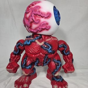 Puede incluir: Una figura grotesca con tem&aacute;tica anat&oacute;mica. La figura presenta una gran cabeza de globo ocular blanco con detalles rosados y rojos en forma de cerebro. El cuerpo es de color rojo intenso con acentos azules en forma de venas, mostrando una forma muscular.