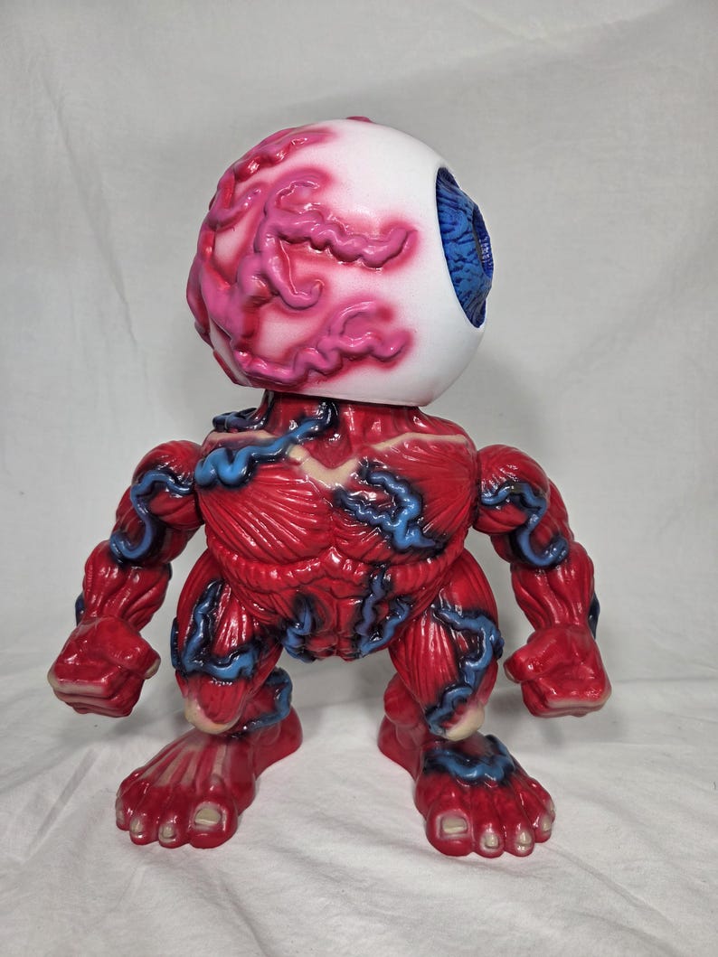 Puede incluir: Una figura grotesca con tem&aacute;tica anat&oacute;mica. La figura presenta una gran cabeza de globo ocular blanco con detalles rosados y rojos en forma de cerebro. El cuerpo es de color rojo intenso con acentos azules en forma de venas, mostrando una forma muscular.
