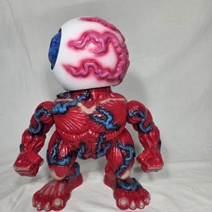 Puede incluir: Una figura de monstruo rojo y azul con una gran cabeza de globo ocular. El globo ocular es blanco con venas rosas y un iris azul. El cuerpo es rojo con venas azules, brazos musculosos y pies.