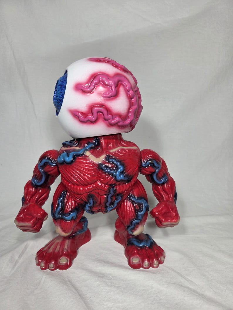 Puede incluir: Una figura de monstruo rojo y azul con una gran cabeza de globo ocular. El globo ocular es blanco con venas rosas y un iris azul. El cuerpo es rojo con venas azules, brazos musculosos y pies.
