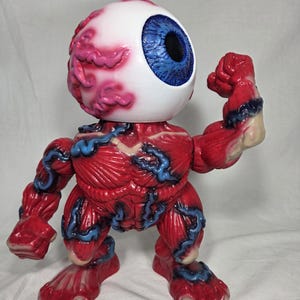 Puede incluir: Una figura anat&oacute;mica roja y azul con una gran cabeza de globo ocular. El globo ocular es blanco con un iris azul y una pupila negra. El cuerpo de la figura es rojo con venas azules y detalles musculares. Est&aacute; flexionando su brazo.