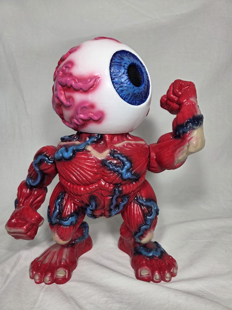 Puede incluir: Una figura anat&oacute;mica roja y azul con una gran cabeza de globo ocular. El globo ocular es blanco con un iris azul y una pupila negra. El cuerpo de la figura es rojo con venas azules y detalles musculares. Est&aacute; flexionando su brazo.