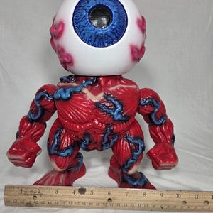 Puede incluir: Una figura estilizada con una cabeza de globo ocular grande, iris azul y escler&oacute;tica blanca. El cuerpo es rojo con detalles azules en forma de venas, mostrando una forma muscular. La figura mide aproximadamente 15 cm de altura.