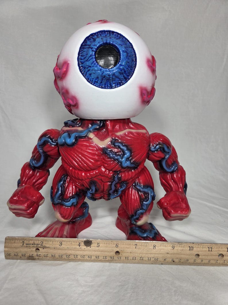 Puede incluir: Una figura estilizada con una cabeza de globo ocular grande, iris azul y escler&oacute;tica blanca. El cuerpo es rojo con detalles azules en forma de venas, mostrando una forma muscular. La figura mide aproximadamente 15 cm de altura.