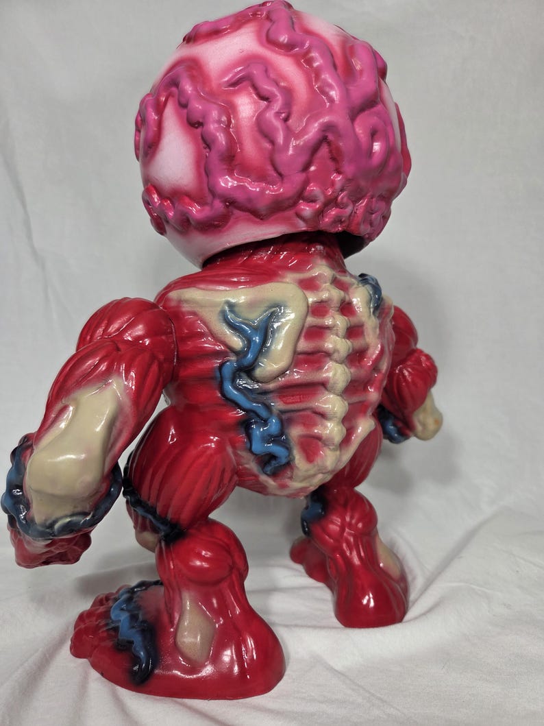 Puede incluir: Figura anat&oacute;mica roja y rosa con m&uacute;sculos, venas y cabeza con forma de cerebro expuestos. El juguete tiene un acabado brillante y una estructura esquel&eacute;tica detallada visible en la espalda. La figura est&aacute; de pie.