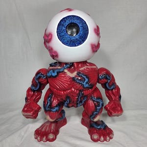 Puede incluir: Una figura de monstruo rojo y azul con una gran cabeza de globo ocular. El globo ocular es blanco con un iris azul y venas rosas. El cuerpo es rojo con venas azules, y la figura tiene brazos, piernas y pies.