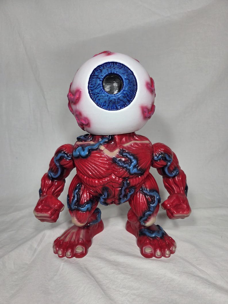 Puede incluir: Una figura de monstruo rojo y azul con una gran cabeza de globo ocular. El globo ocular es blanco con un iris azul y venas rosas. El cuerpo es rojo con venas azules, y la figura tiene brazos, piernas y pies.