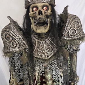 May include: A skeletal figure wearing a detailed crown and armor. The figure has glowing red eyes and long black hair. The armor includes shoulder pads, a chest piece, and chainmail. The overall design suggests a fantasy or Halloween theme.