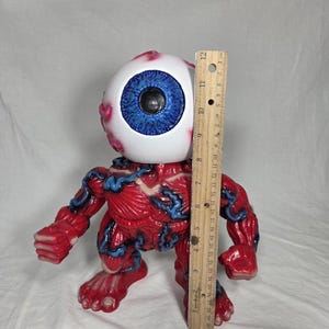 Puede incluir: Una figura anat&oacute;mica roja y azul con una gran cabeza de globo ocular. La figura mide aproximadamente 25 cm de altura, con una representaci&oacute;n detallada de m&uacute;sculos y venas. El globo ocular tiene un iris azul y una pupila negra.