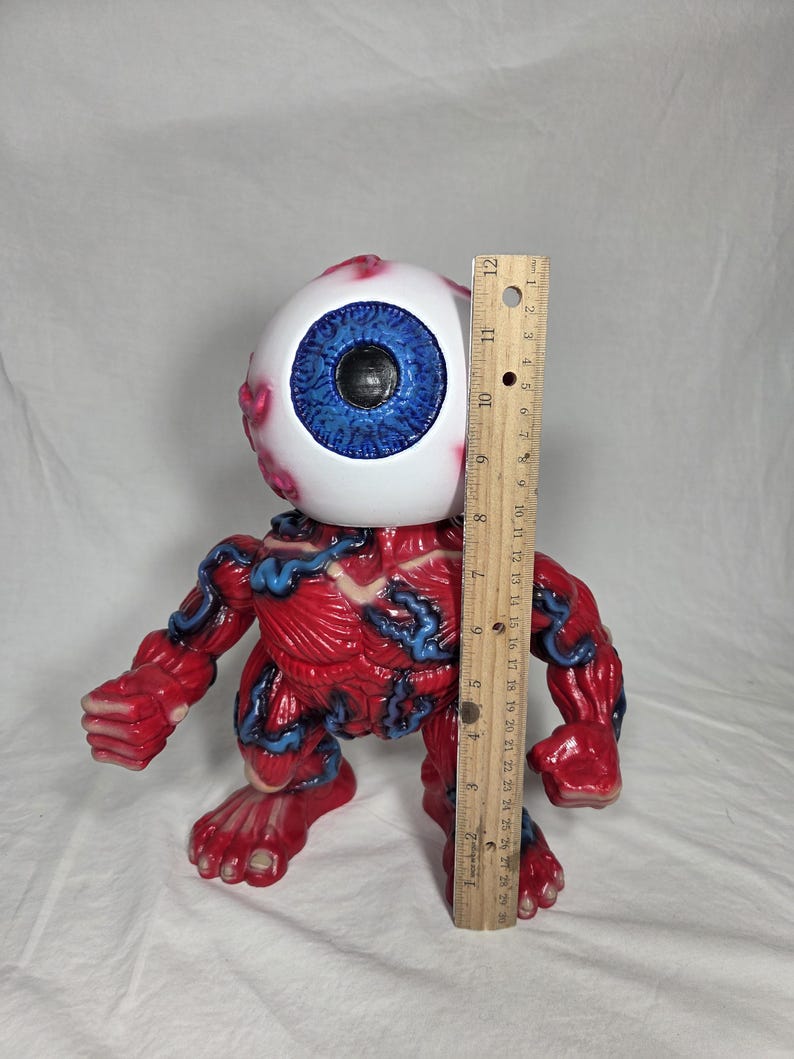Puede incluir: Una figura anat&oacute;mica roja y azul con una gran cabeza de globo ocular. La figura mide aproximadamente 25 cm de altura, con una representaci&oacute;n detallada de m&uacute;sculos y venas. El globo ocular tiene un iris azul y una pupila negra.