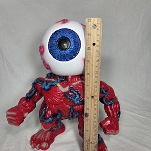 Puede incluir: Una figura anat&oacute;mica roja y azul con una gran cabeza de globo ocular. La figura mide aproximadamente 25 cm de altura, medida con una regla de madera. El globo ocular tiene un iris azul y una pupila negra.
