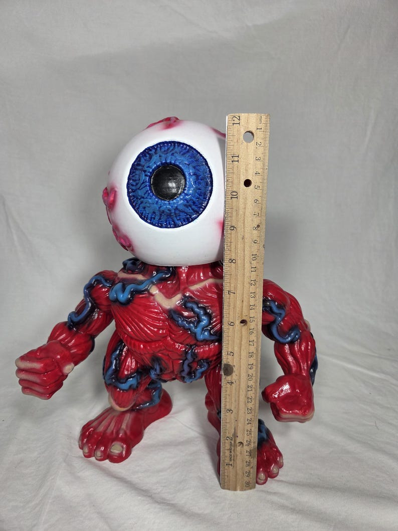 Puede incluir: Una figura anat&oacute;mica roja y azul con una gran cabeza de globo ocular. La figura mide aproximadamente 25 cm de altura, medida con una regla de madera. El globo ocular tiene un iris azul y una pupila negra.