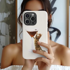 May include: A white smartphone case featuring a graphic illustration of a brown tabby cat reaching up toward a martini glass filled with a dark liquid and topped with three coffee beans. The case has a black camera lens cutout at the top.