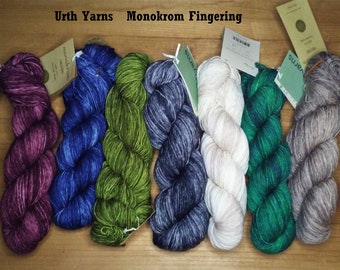 May include: A collection of Urth Yarns Monokrom Fingering yarn skeins in various colors, including purple, blue, green, white, and gray. Each skein is labeled with a tag. The yarns are arranged on a wooden surface.