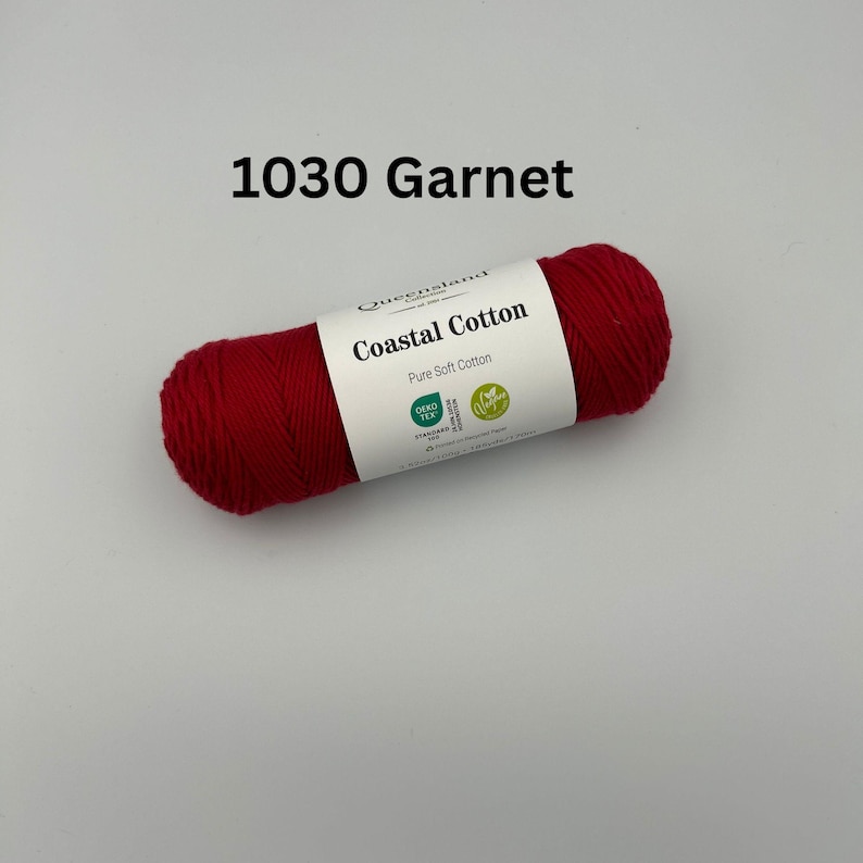 K&ouml;nnte beinhalten: Eine rote Garnrolle mit der Aufschrift "1030 Garnet" und "Coastal Cotton", mit dem Text "Pure Soft Cotton" und den Logos "Oeko-Tex Standard 100" und "GOTS" auf dem Etikett.