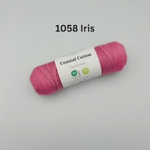 K&ouml;nnte beinhalten: Eine rosa Garnrolle von Coastal Cotton mit einem wei&szlig;en Etikett, auf dem "Coastal Cotton Pure Soft Cotton" steht und ein gr&uuml;nes Oeko-Tex Standard 100 Logo und ein gr&uuml;nes Vegan Logo abgebildet sind. Das Garn ist mit 1058 Iris gekennzeichnet.