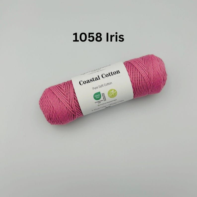 K&ouml;nnte beinhalten: Eine rosa Garnrolle von Coastal Cotton mit einem wei&szlig;en Etikett, auf dem "Coastal Cotton Pure Soft Cotton" steht und ein gr&uuml;nes Oeko-Tex Standard 100 Logo und ein gr&uuml;nes Vegan Logo abgebildet sind. Das Garn ist mit 1058 Iris gekennzeichnet.