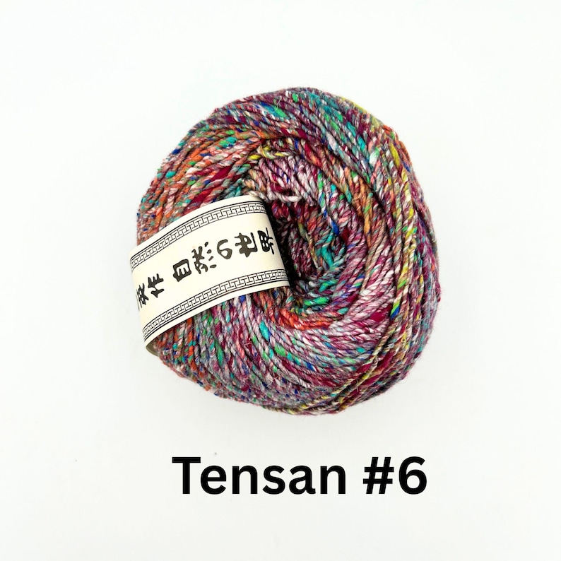 K&ouml;nnte beinhalten: Ein Kn&auml;uel bunter Wolle mit einem Etikett mit japanischem Text. Die Wolle ist eine Mischung aus Rot, Lila, Gr&uuml;n und Orange. Der Text "Tensa #6" ist darunter in fetter Schrift gedruckt.