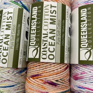 May include: Two skeins of yarn in a speckled pattern. The yarn is labeled "Coastal Cotton Ocean Mist" and is 100% cotton. The skein on the left is white with blue, green, and red speckles. The skein on the right is a light peach color with orange, yellow, pink, and purple speckles.