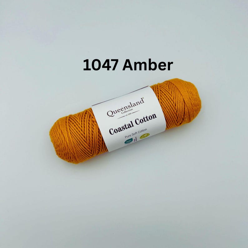 K&ouml;nnte beinhalten: Ein Kn&auml;uel amberfarbenes Garn mit der Aufschrift "Queensland Collection Coastal Cotton Pure Soft Cotton". Das Garn ist 1047 Amber.