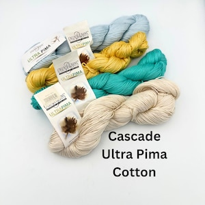May include: A collection of Cascade Ultra Pima Cotton yarn in various colors, including light blue, yellow, teal, and cream. The yarn is displayed with its labels, showcasing the brand and cotton type. The image is well-lit, highlighting the texture and colors of the yarn.
