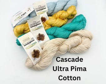 May include: A collection of Cascade Ultra Pima Cotton yarn in various colors, including light blue, yellow, teal, and cream. The yarn is displayed with its labels, showcasing the brand and cotton type. The image is well-lit, highlighting the texture and colors of the yarn.