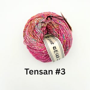 K&ouml;nnte beinhalten: Ein Kn&auml;uel leuchtend rosa Garn mit gelben, blauen und wei&szlig;en Sprenkeln. Ein Etikett mit japanischem Text ist um das Garn gewickelt. Der Text "Tensa #3" ist unten im Bild in Schwarz gedruckt.