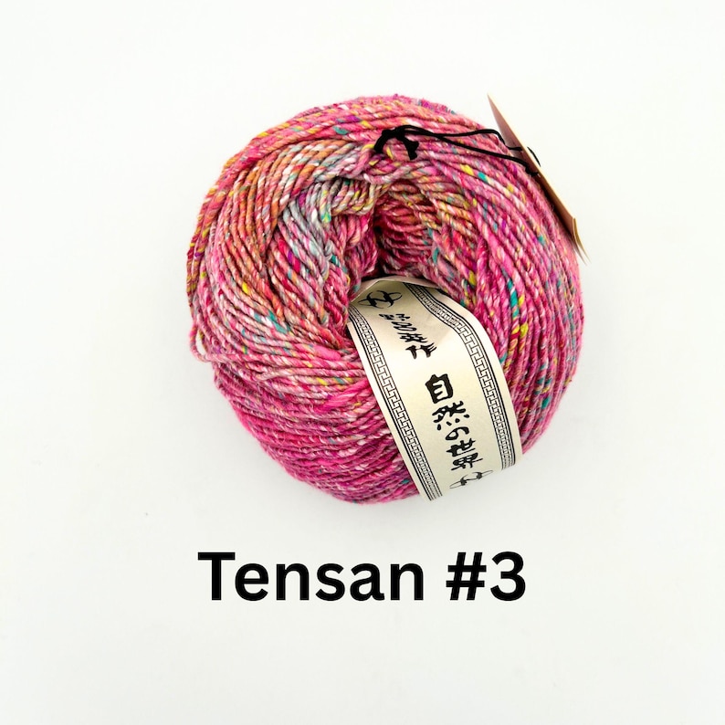 K&ouml;nnte beinhalten: Ein Kn&auml;uel leuchtend rosa Garn mit gelben, blauen und wei&szlig;en Sprenkeln. Ein Etikett mit japanischem Text ist um das Garn gewickelt. Der Text "Tensa #3" ist unten im Bild in Schwarz gedruckt.