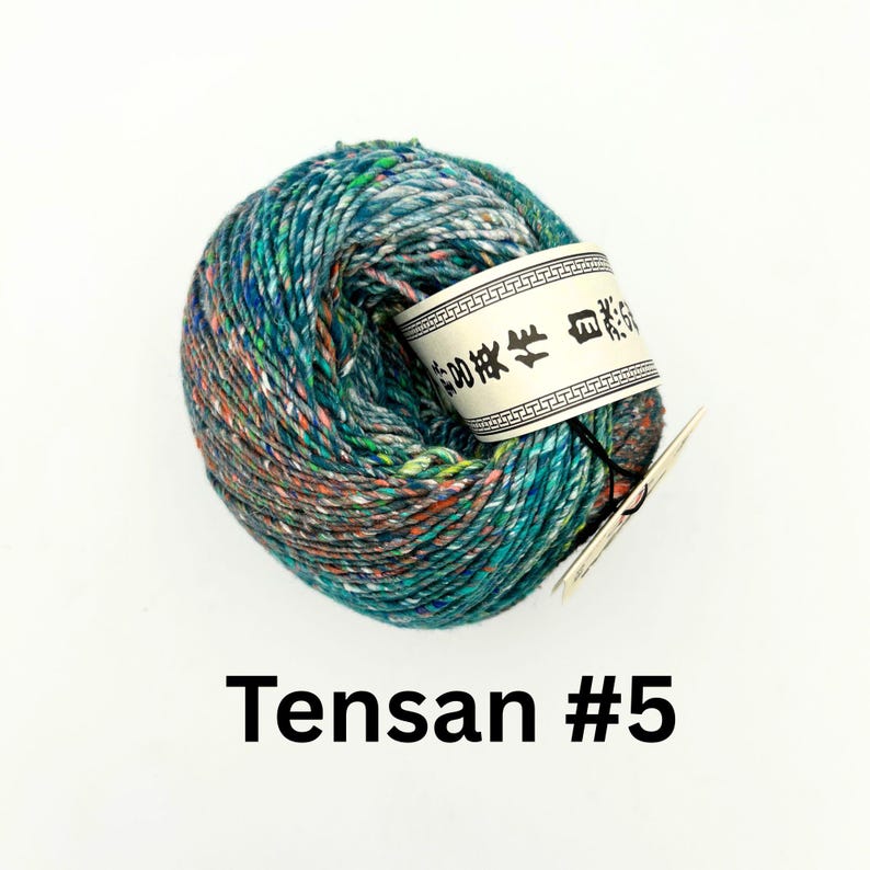 K&ouml;nnte beinhalten: Ein Kn&auml;uel Tensa #5 Garn in T&uuml;rkis-, Blau- und Oranget&ouml;nen. Das Garn hat ein gesprenkeltes Aussehen und ein wei&szlig;es Etikett mit asiatischen Schriftzeichen. Der Text "Tensa #5" ist unten im Bild in Schwarz gedruckt.