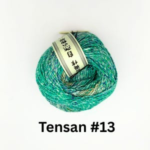 K&ouml;nnte beinhalten: Ein Kn&auml;uel gr&uuml;n-blau gesprenkelter Wolle mit einem wei&szlig;en Etikett mit japanischem Text. Das Garn ist mit "Tensan #13" beschriftet. Das Garn ist fest gewickelt und wirkt weich.