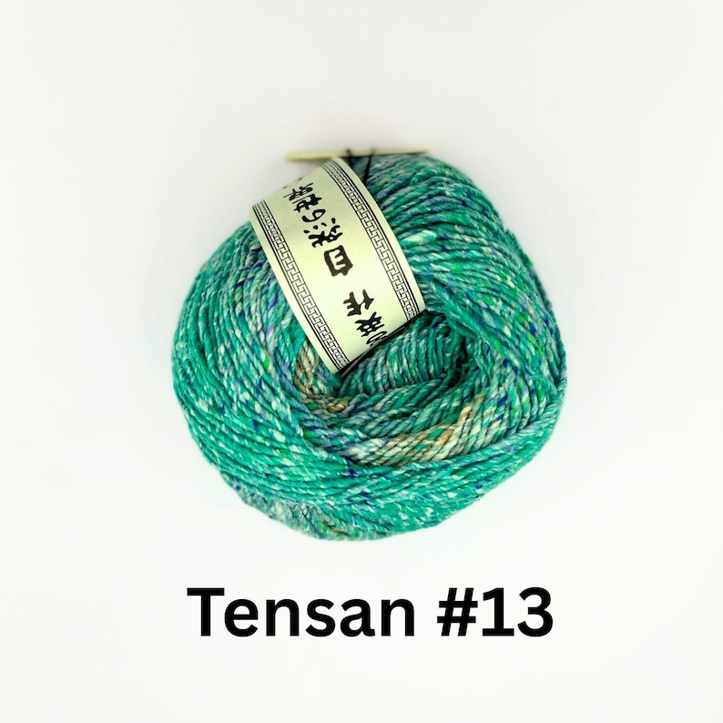 K&ouml;nnte beinhalten: Ein Kn&auml;uel gr&uuml;n-blau gesprenkelter Wolle mit einem wei&szlig;en Etikett mit japanischem Text. Das Garn ist mit "Tensan #13" beschriftet. Das Garn ist fest gewickelt und wirkt weich.