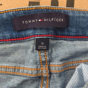 May include: Close-up of blue denim jeans with the Tommy Hilfiger label visible. The label is navy blue with the brand name in white and a red and white logo. The jeans have a size 10 tag and are made in Vietnam.