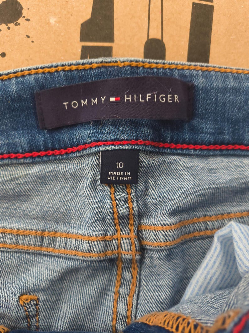 May include: Close-up of blue denim jeans with the Tommy Hilfiger label visible. The label is navy blue with the brand name in white and a red and white logo. The jeans have a size 10 tag and are made in Vietnam.