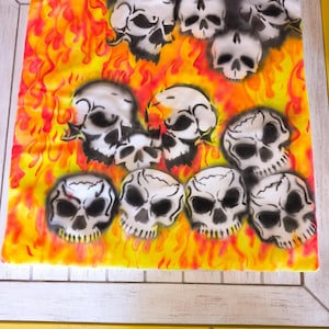 May include: A decorative square fabric featuring a design of white skulls against a backdrop of flames in shades of yellow, orange, and red. The skulls have black shading, and the overall effect is a fiery, gothic theme.