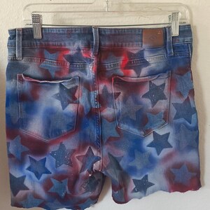 May include: Denim shorts with a patriotic design. The shorts feature a blue wash with red and blue star patterns. The shorts are hanging on a clear plastic hanger.