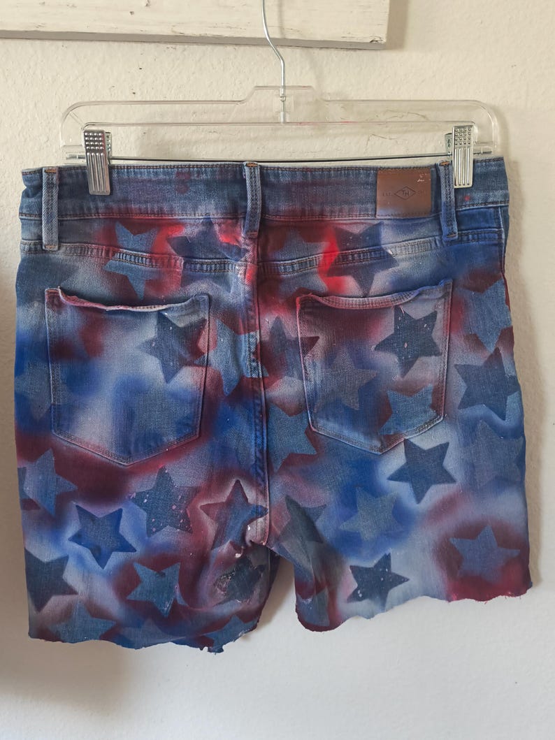 May include: Denim shorts with a patriotic design. The shorts feature a blue wash with red and blue star patterns. The shorts are hanging on a clear plastic hanger.
