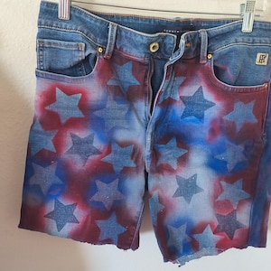 May include: Denim shorts with a patriotic design. The shorts feature a blue denim base with a pattern of red and blue stars. The shorts have a zipper fly, pockets, and a raw hem.