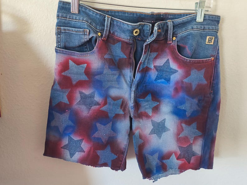 May include: Denim shorts with a patriotic design. The shorts feature a blue denim base with a pattern of red and blue stars. The shorts have a zipper fly, pockets, and a raw hem.