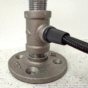 May include: A dark gray metal pipe fitting base with a circular flange featuring four screw holes. A vertical threaded pipe segment connects to a T-shaped fitting, which holds a black braided electrical cord extending to the right side.