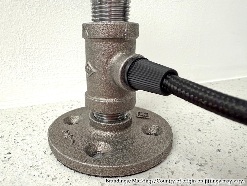 May include: A dark gray metal pipe fitting base with a circular flange featuring four screw holes. A vertical threaded pipe segment connects to a T-shaped fitting, which holds a black braided electrical cord extending to the right side.