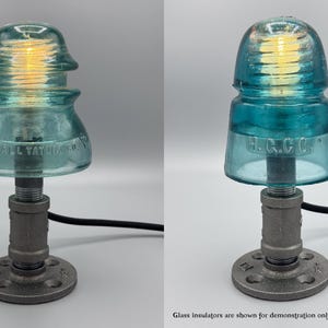 May include: A table lamp featuring a vintage teal glass electrical insulator as a shade, mounted on a dark gray industrial metal pipe base with a circular flange. The lamp includes a warm glowing light bulb and a black power cord.
