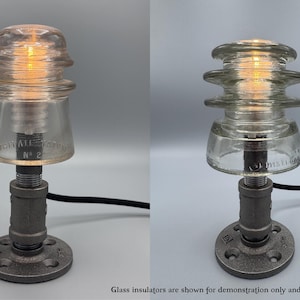May include: A table lamp base constructed from dark gray industrial metal piping with a circular floor flange, a vertical pipe stem, and a threaded socket top. The lamp features a black power cord and is shown with a clear glass insulator.