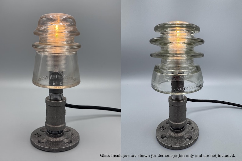 May include: A table lamp base constructed from dark gray industrial metal piping with a circular floor flange, a vertical pipe stem, and a threaded socket top. The lamp features a black power cord and is shown with a clear glass insulator.