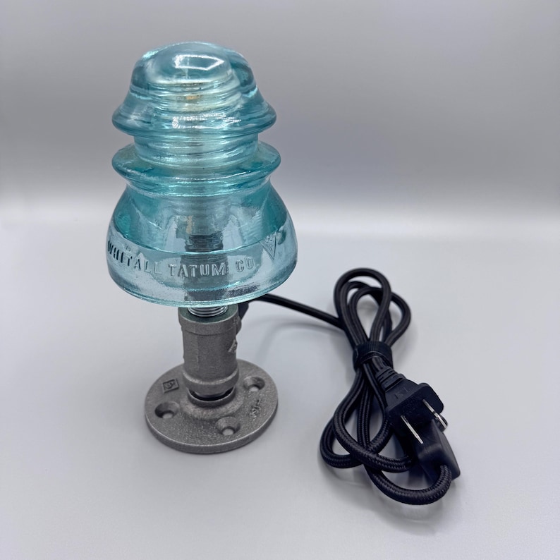 May include: A table lamp featuring a vintage light blue glass electrical insulator marked Whitall Tatum Co. mounted on a dark gray industrial metal pipe base with a circular flange and a black electrical cord with a standard two-prong plug.