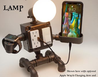 May include: A dark metal robot-shaped lamp with a round white bulb head, a central electrical outlet and switch, and two arms. One arm holds a smartphone, while the other features a charging dock for a smartwatch. Text reads ROBOT LAMP.