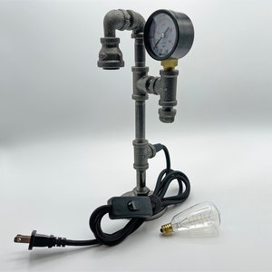 May include: A table lamp constructed from dark gray industrial metal pipes featuring a circular pressure gauge with a white face and black markings. The lamp includes a black power cord with an inline switch and a separate clear glass light bulb.