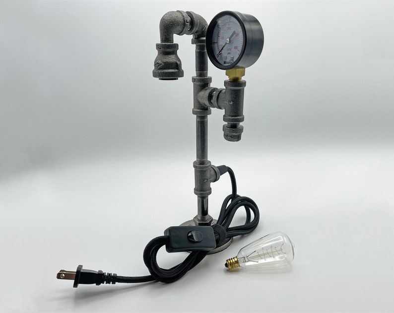 May include: A table lamp constructed from dark gray industrial metal pipes featuring a circular pressure gauge with a white face and black markings. The lamp includes a black power cord with an inline switch and a separate clear glass light bulb.