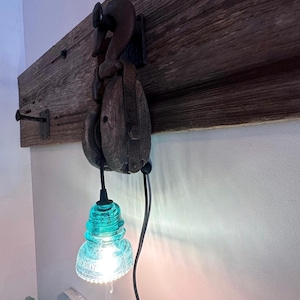 May include: A wall-mounted light fitting featuring a weathered wooden board with a dark metal hook and a vintage wooden pulley block. A bright, translucent turquoise glass insulator serves as the lampshade, suspended by a black electrical cord.